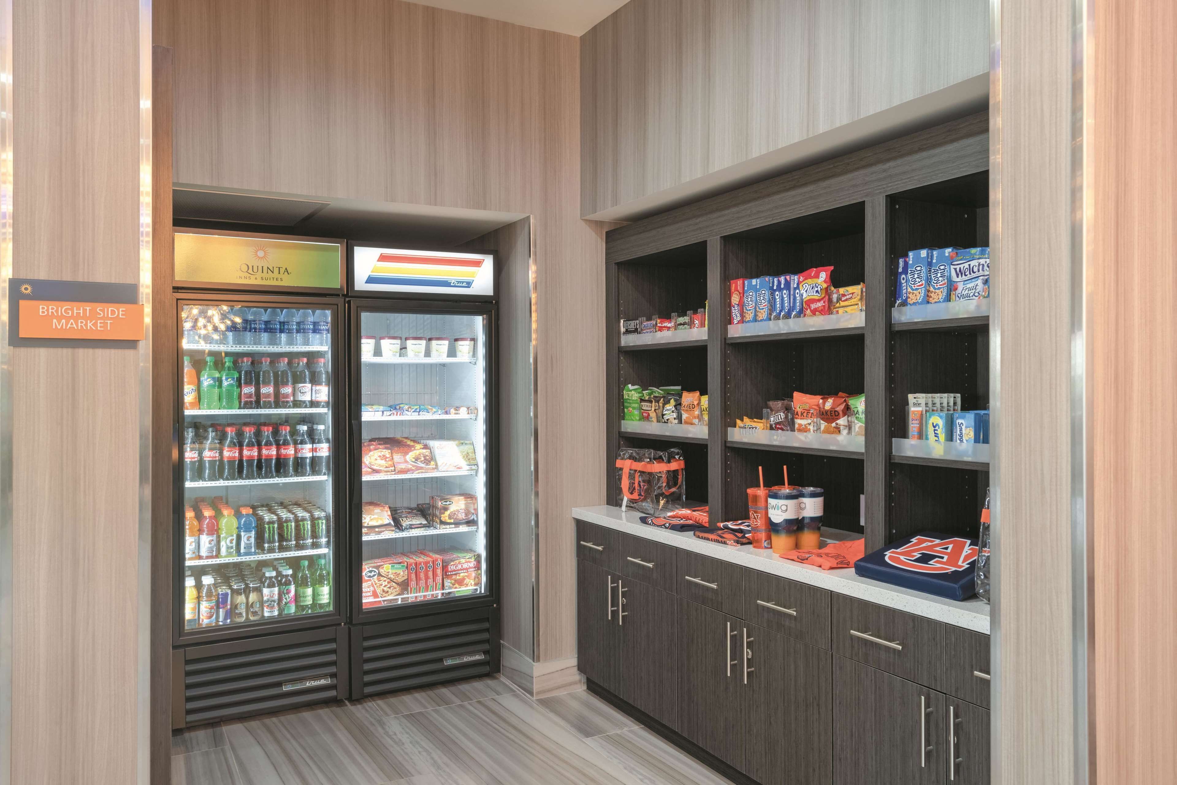 A minibar area with a refrigerator stocked with beverages and snacks, a shelf with packaged food items, and a sign indicating the location of the minibar.