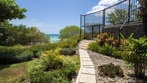 Ein Weg führt zu einem Balkon mit Blick auf das Meer, umgeben von tropischen Pflanzen und einer Steinmauer.