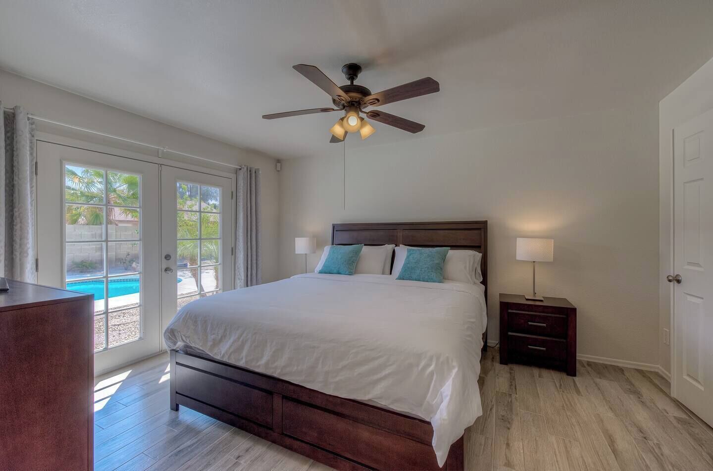 A bedroom with a large bed, a ceiling fan, a nightstand with a lamp, and a view of a swimming pool through a sliding glass door.
