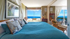Une chambre avec un grand lit, une vue sur l’océan, un fauteuil en osier et une petite table avec une lampe.