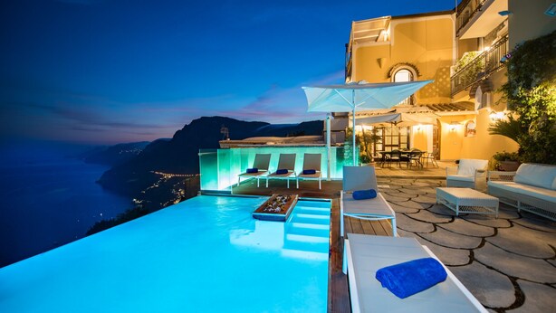 Uma piscina infinita com espreguiçadeiras e uma mesa, com vista para uma paisagem montanhosa.