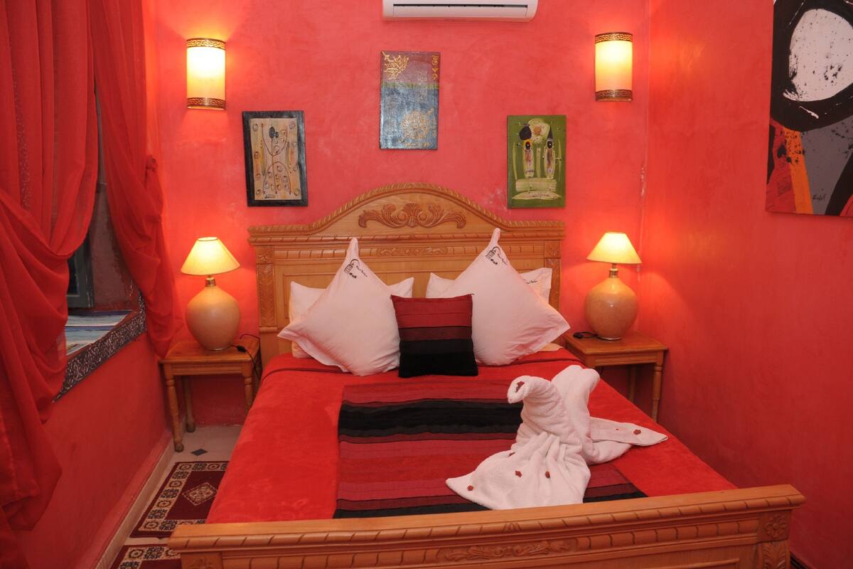 Habitación de hotel con cama de paredes rojas, dos lámparas de noche y obras de arte en las paredes.