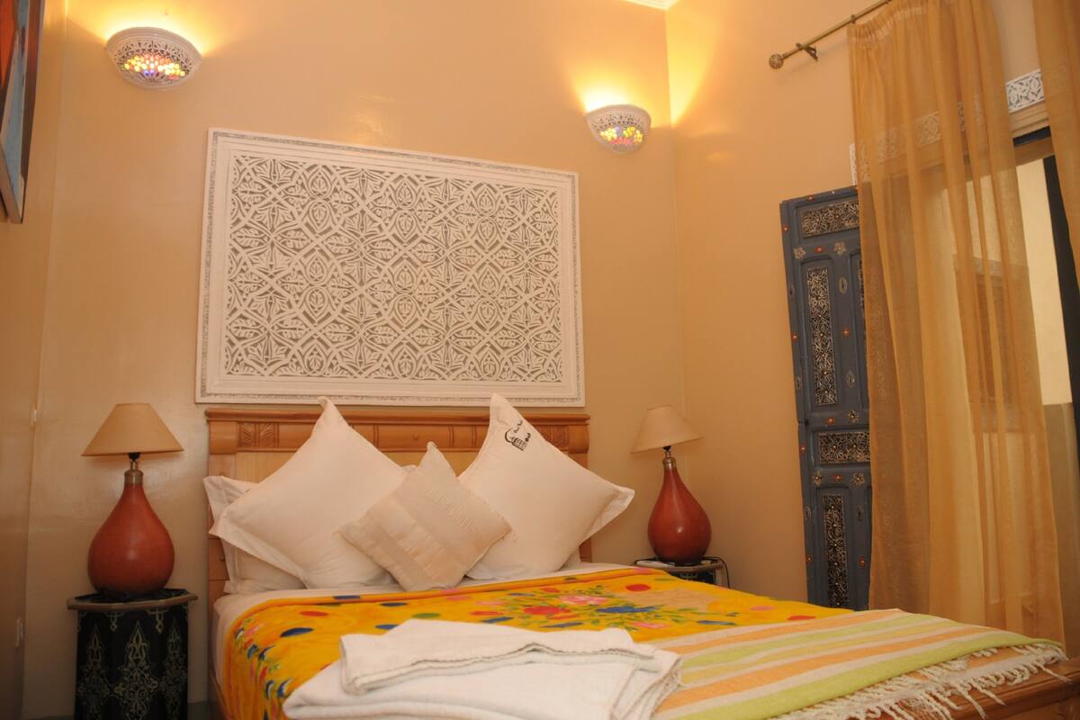 Habitación de hotel con cama, dos lámparas de noche, un tapiz decorativo y una puerta azul con diseños intrincados.
