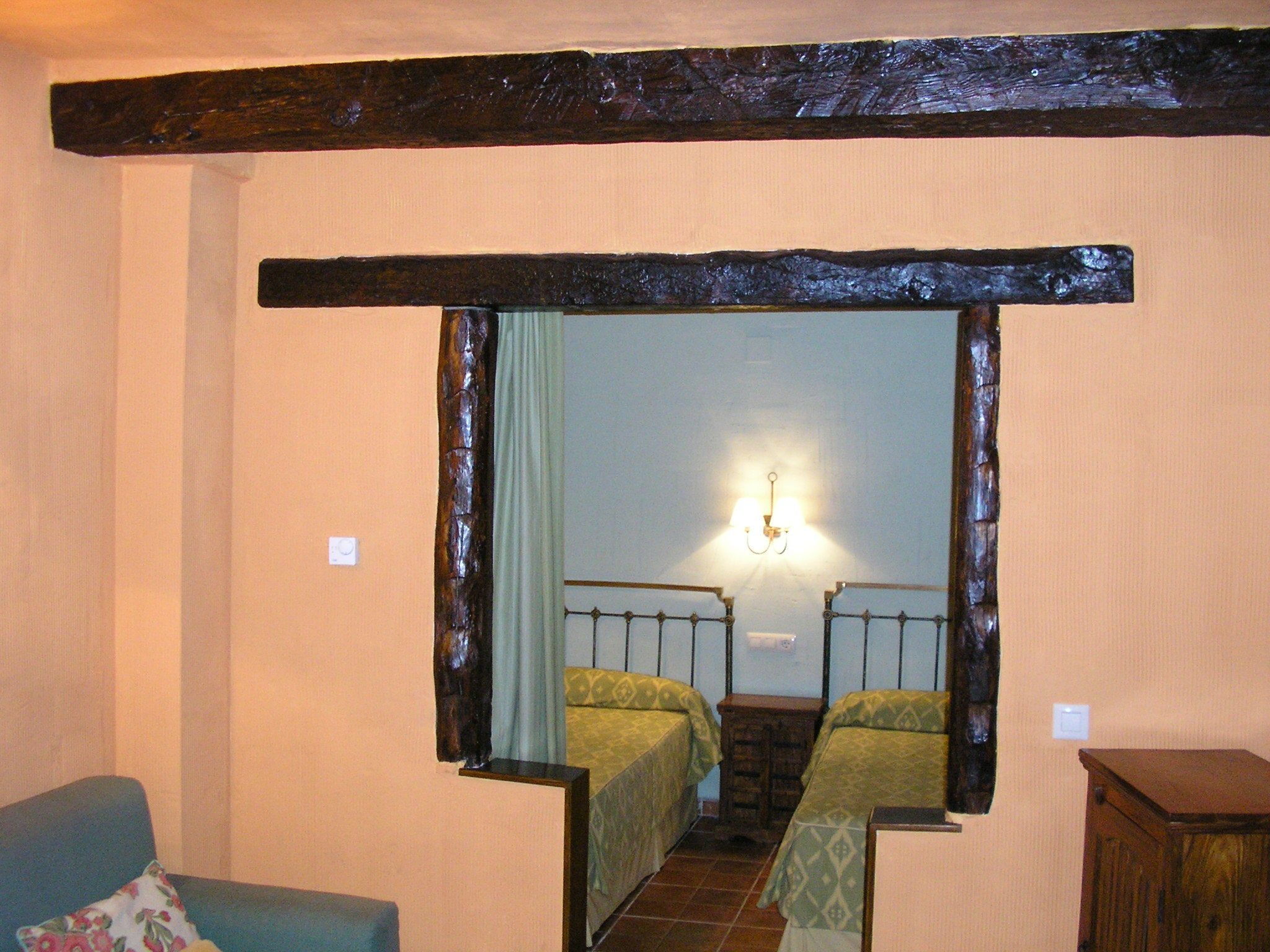 A room with a wooden ceiling beam, a mirror reflecting a staircase, two beds with green bedding, a small wooden cabinet, and a blue chair with a floral pillow.
