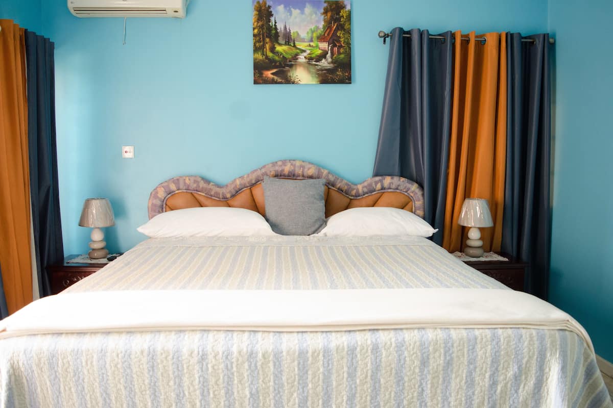Une chambre à coucher comprenant un lit, deux tables de chevet, un climatiseur fixé au mur et un tableau représentant un paysage.