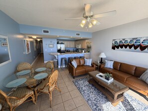 Sala de estar com sofá marrom, mesa de jantar de vidro e cadeiras de vime, cozinha ao fundo e um ventilador de teto.