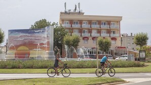 A hotel with a large mural, a fountain, and a road with cyclists. - Hotel Silvia (Castello d'Empuries)