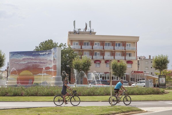 A hotel with a large mural, a fountain, and a road with cyclists. - Hotel Silvia (Castello d'Empuries)