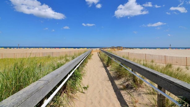 A wooden boardwalk leading to a sandy beach with a clear blue sky and scattered clouds.