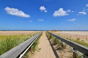 Ein hölzerner Steg führt zu einem Sandstrand, der von einem klaren, blauen Himmel mit vereinzelten Wolken gesäumt wird.