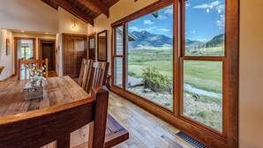 A dining area with a wooden table and chairs, large windows with a mountain view, and wooden beams on the ceiling.