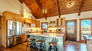 A kitchen with wooden cabinets, a stainless steel refrigerator, a large island with stools, and a view of the outdoors through a door.