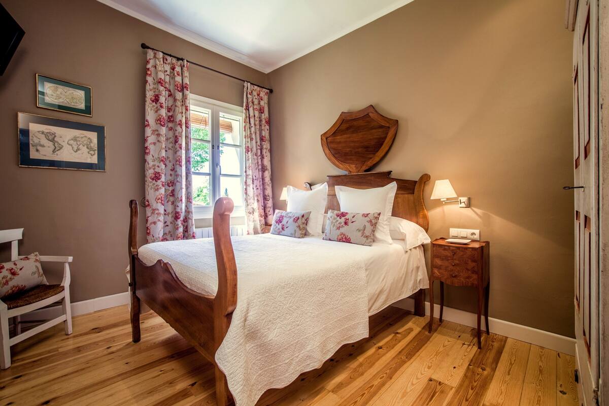 Un dormitorio con cama de madera, cortinas florales, una mesita de noche con lámpara y cuadros enmarcados en la pared.
