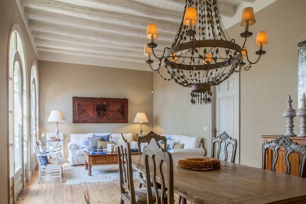 A dining area with a wooden table, chairs, and a chandelier. There is a sofa and a lamp in the background.