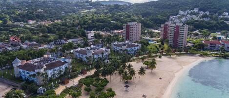 An aerial view of a coastal resort with beachfront villas, a sandy beach, and a backdrop of lush hills.