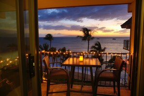 A balcony with a table and chairs, overlooking the ocean with a sunset view.