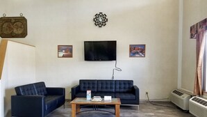 Un salon avec un canapé bleu foncé, un fauteuil plus petit et une table basse en bois. Un téléviseur à écran plat est fixé au mur, et deux climatiseurs sont posés sur le sol.