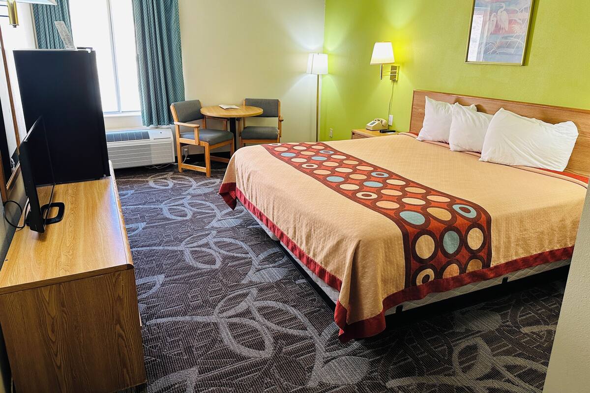 A hotel room with a bed, a desk with a computer, a chair, a small table, and a lamp.
