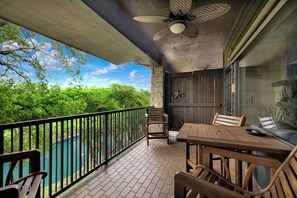 A balcony with a view of a river, a wooden table and chairs, a ceiling fan, and a stone wall.