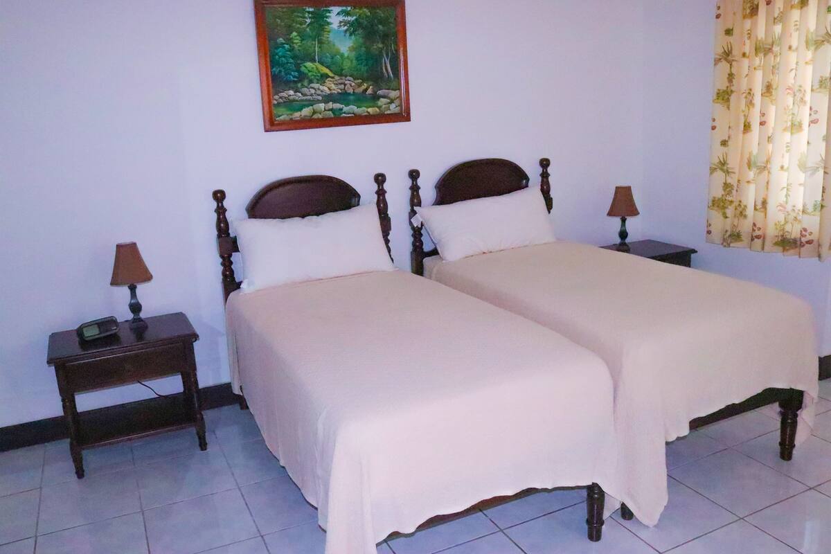 A double bed with a wooden frame and white bedding, two bedside tables with lamps, and a painting of a forest scene on the wall.