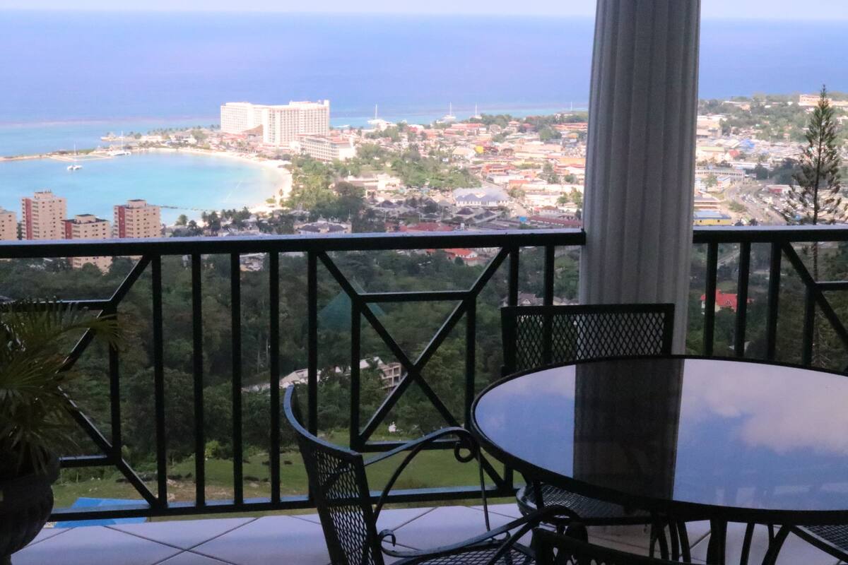 A balcony with a table and chairs, overlooking a coastal cityscape with buildings and a beach.