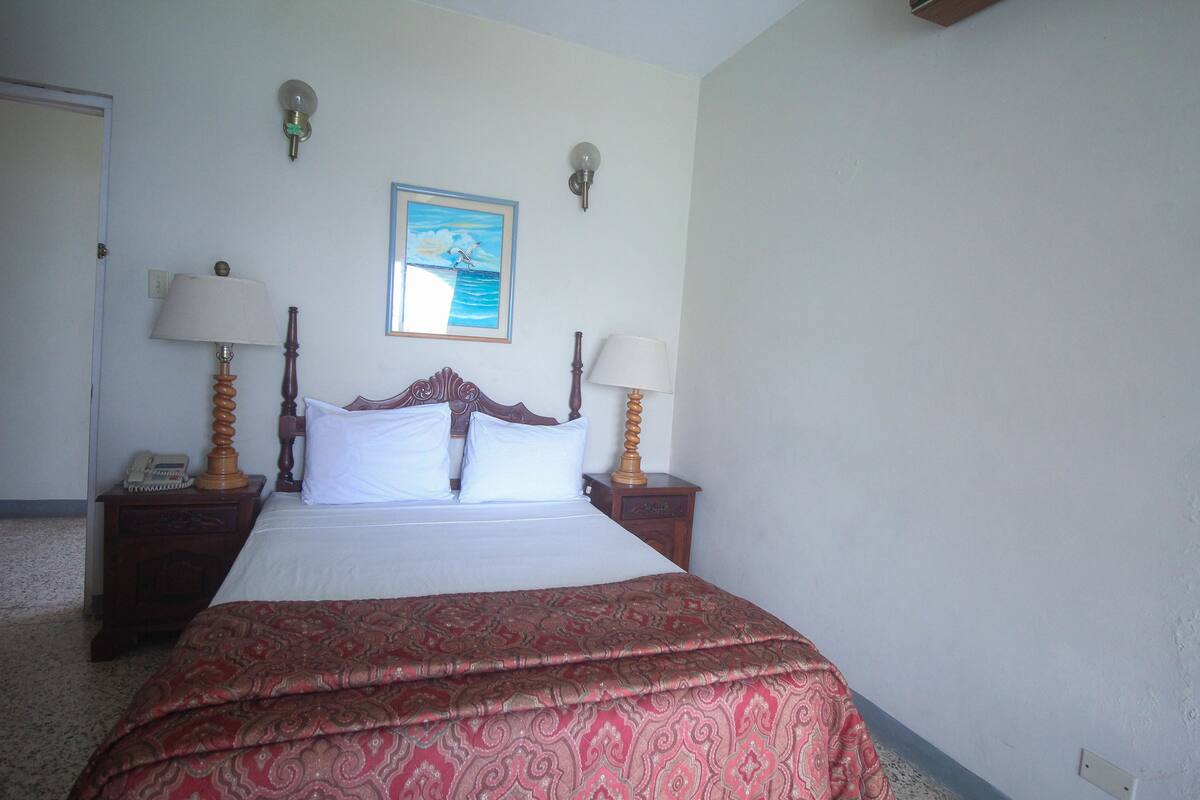 A neatly made bed with a patterned bedspread, two bedside lamps, a nightstand with a telephone, and a framed picture on the wall.