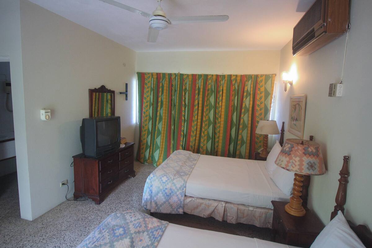 A hotel room with two beds, a television, a ceiling fan, and a window with curtains.