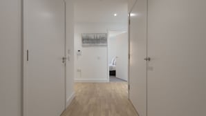 A hallway with white walls and wooden flooring leading to a bedroom with a bed and a painting on the wall. - Fortified Apartment (Chelmsford)