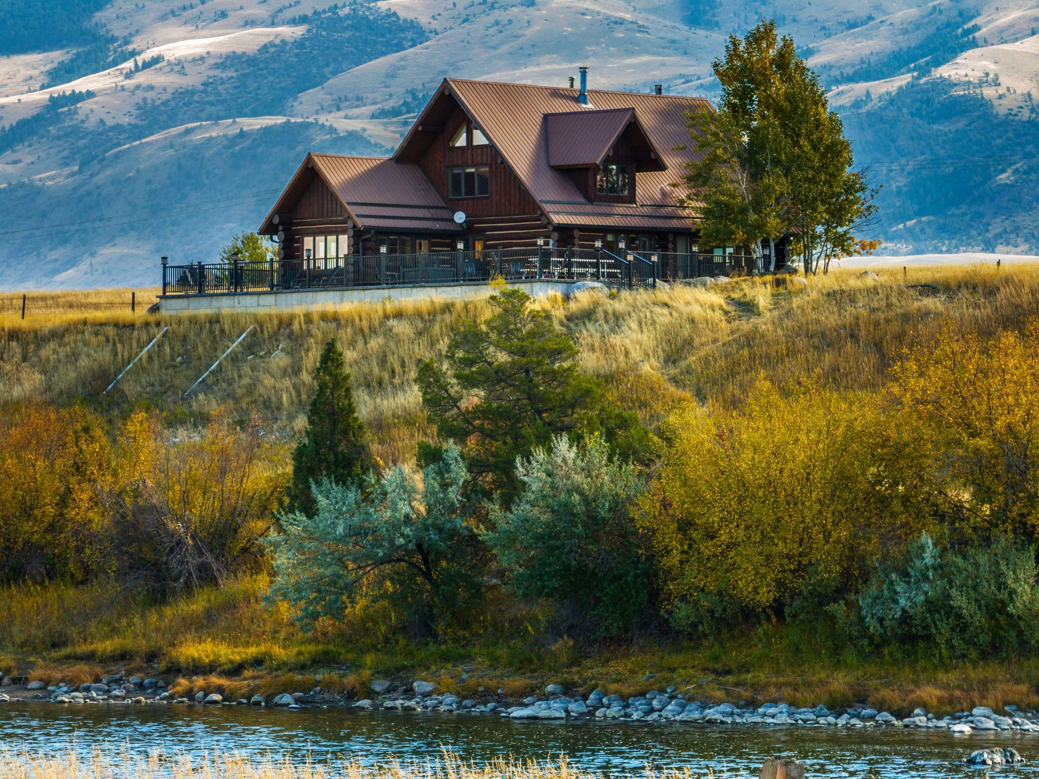 A log cabin with a metal roof, situated on a hill overlooking a river and mountains.