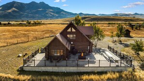 Une cabane en rondins dotée d’un patio, entourée d’une clôture, et située dans un cadre magnifique avec des montagnes et des champs en arrière-plan.
