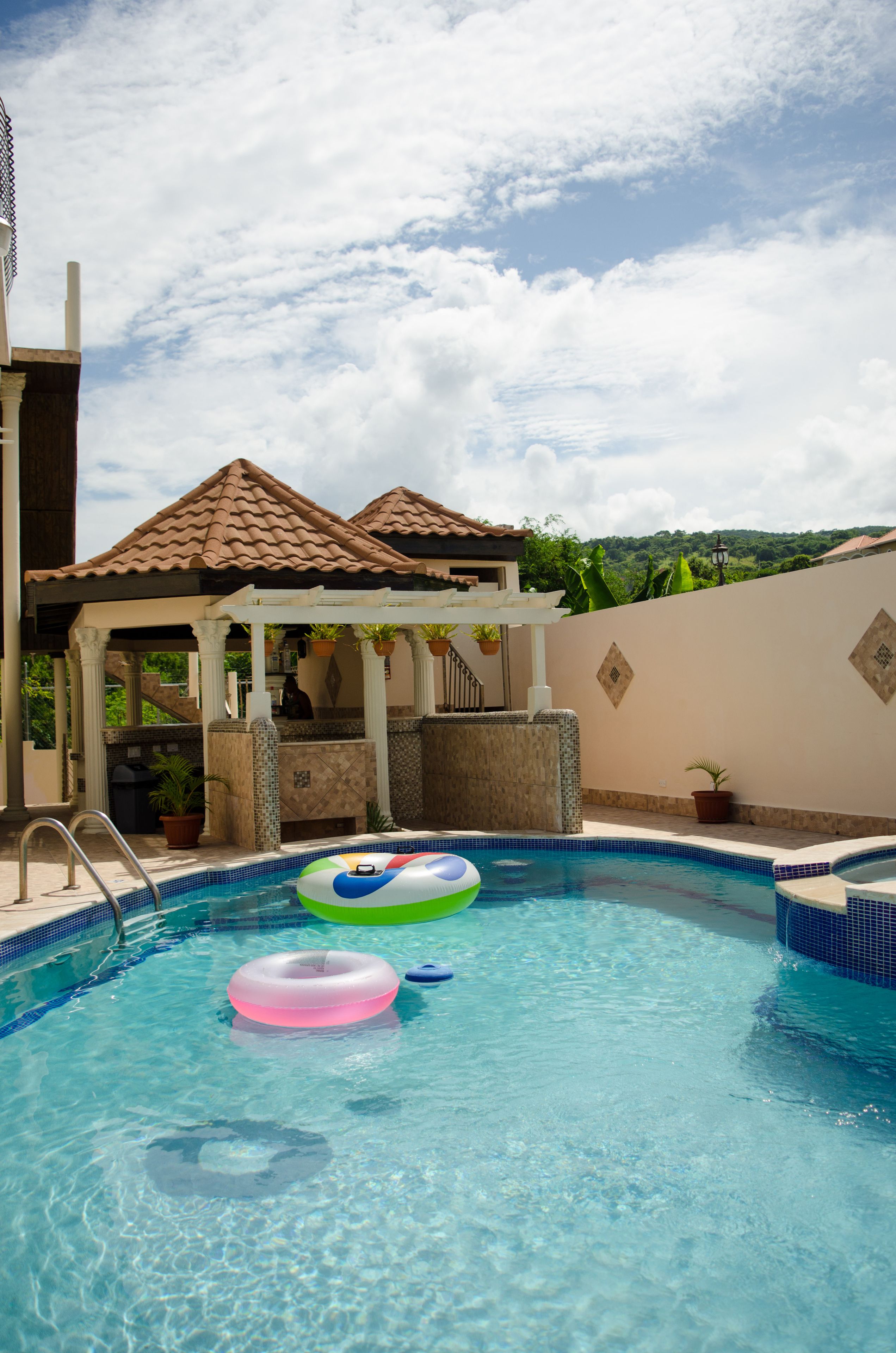 A swimming pool with two inflatable rings, a covered seating area, and a view of greenery in the background.
