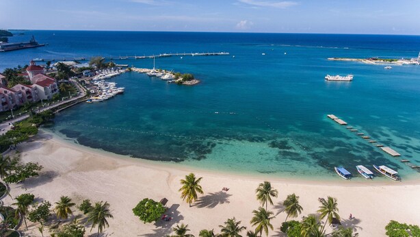 An aerial view of a coastal area with a sandy beach, clear blue waters, and a marina with boats. - Conch Shell Studio at Sandcastles (Ocho Rios)