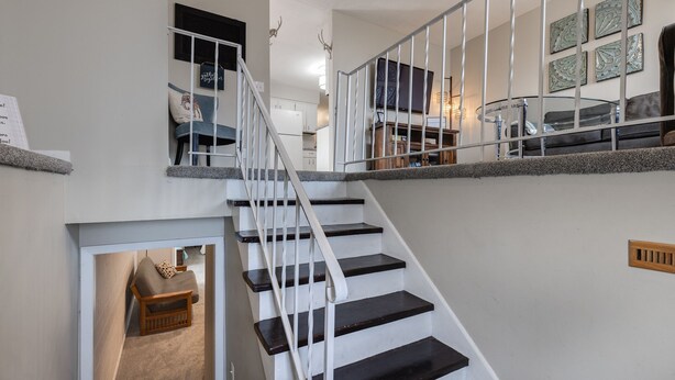 Eine Treppe mit weißen Geländern führt zu einem Wohnbereich mit Fernseher, Glastisch und Sofa.