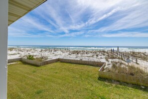 Blick von einer überdachten Veranda auf einen Strand, mit einem Holzzaun und einer grünen Fläche im Vordergrund.