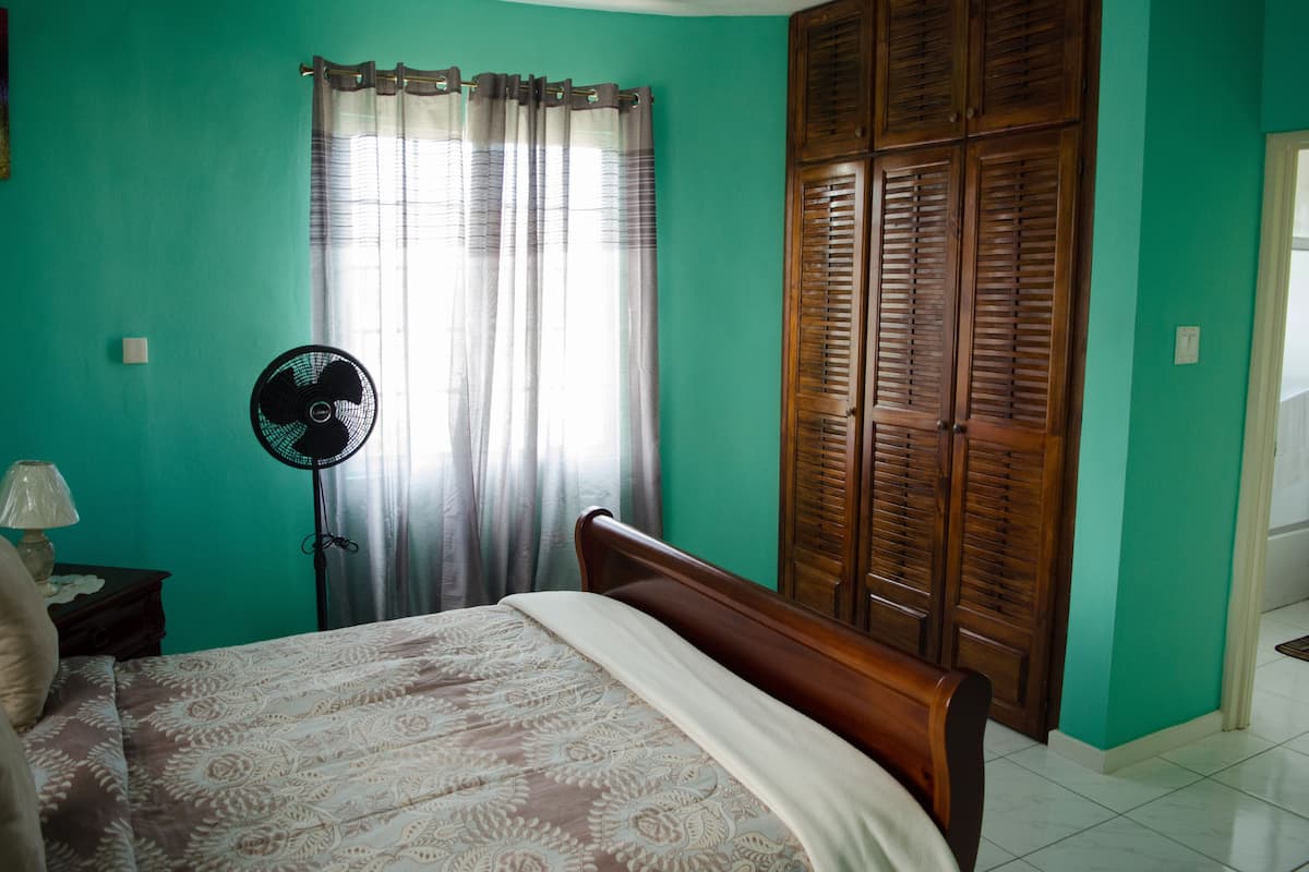 A bedroom with a bed, a standing fan, a nightstand, and a wooden wardrobe.