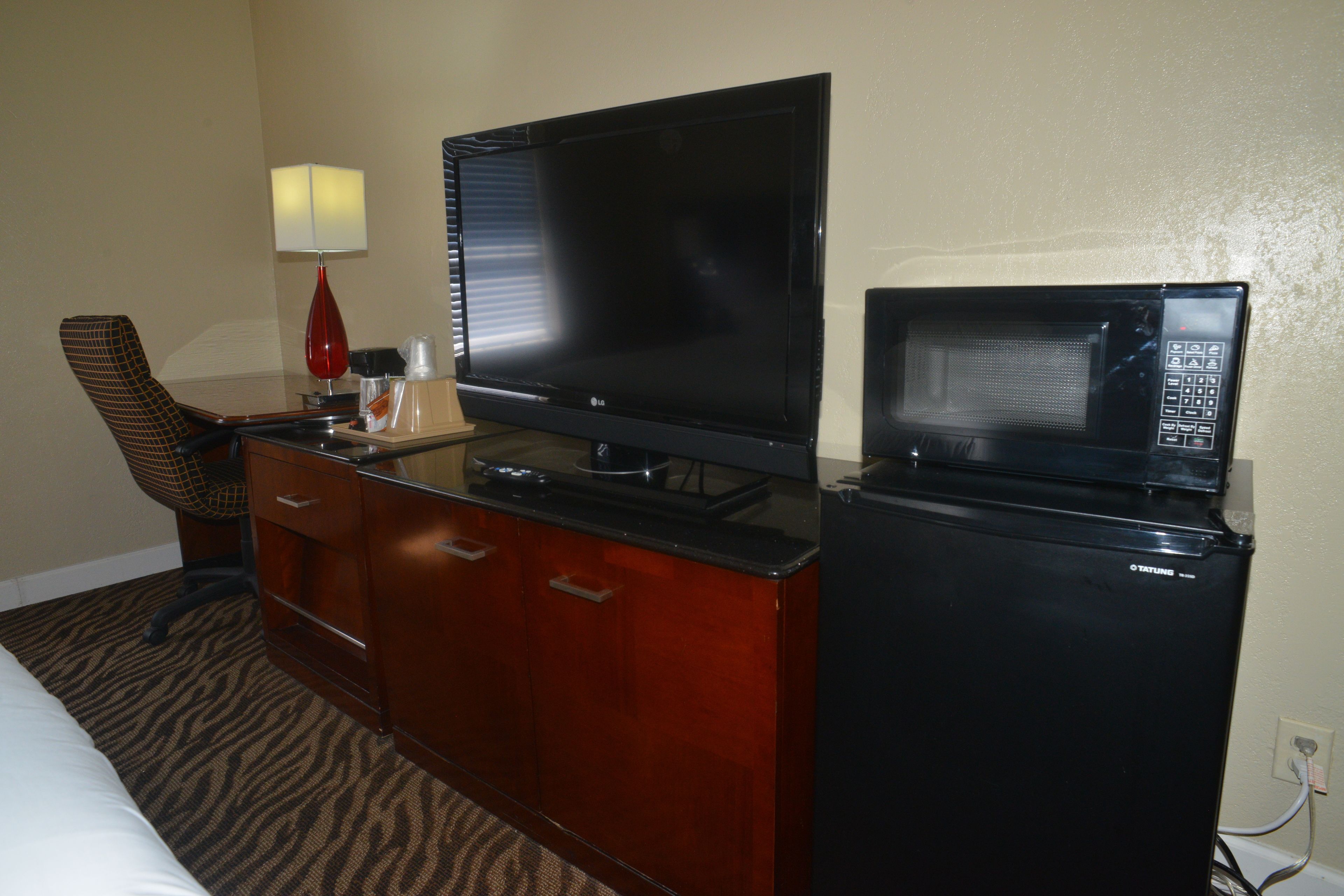 A hotel room with a bed, a TV, a microwave, a desk with a lamp, and a chair.