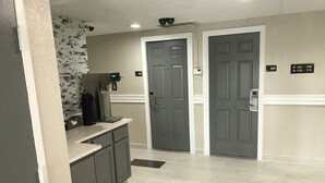A hotel lobby with a coffee machine, a trash bin, and two grey doors with numbered signs.