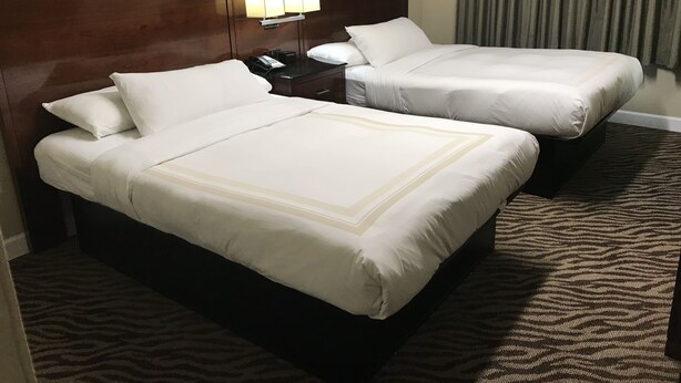 A hotel room with two beds, a wooden headboard, a bedside table with a lamp, and a patterned carpet.