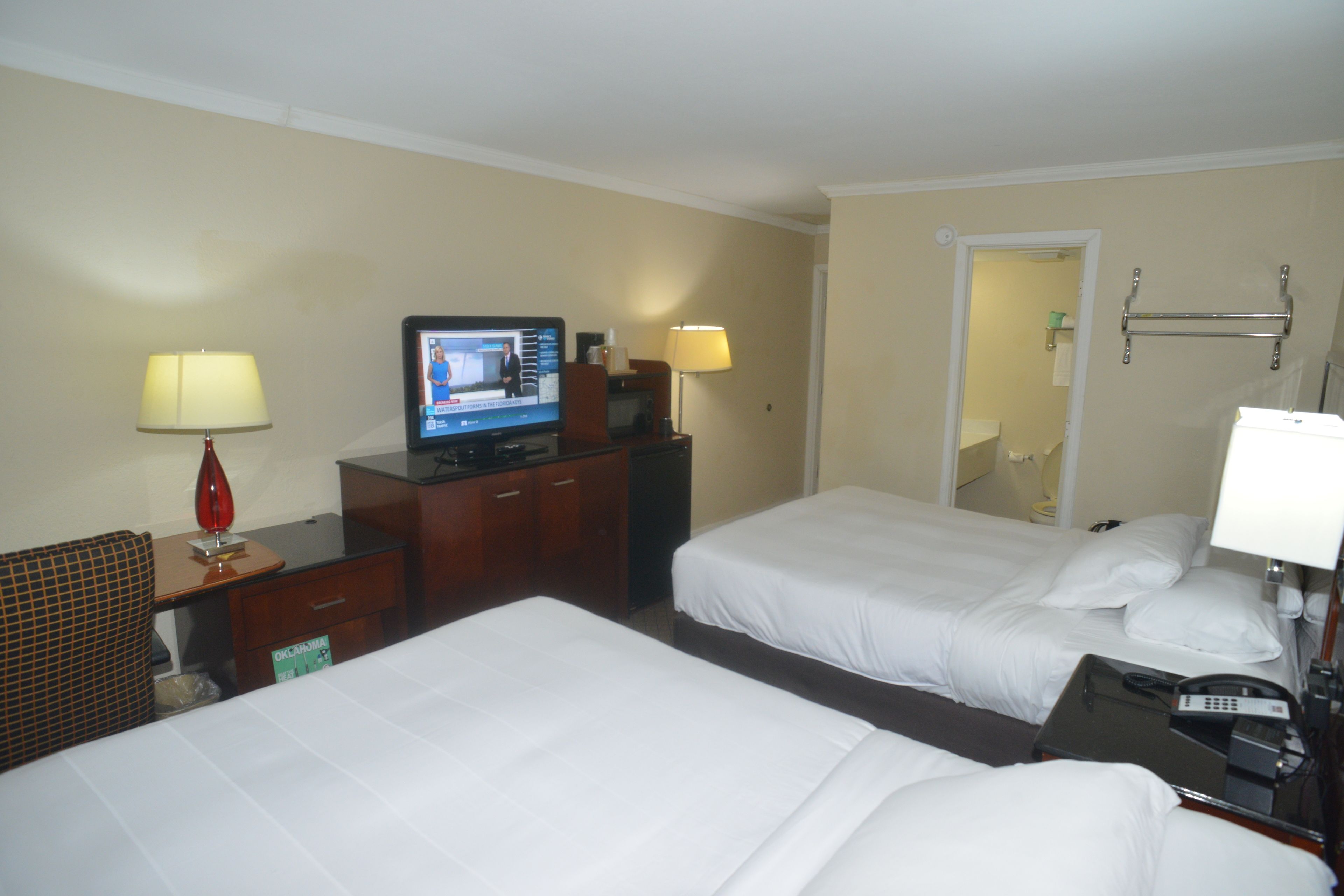 Ein Hotelzimmer mit zwei Betten, einem Fernseher, einem Schreibtisch mit Lampe und einem sichtbaren Badezimmer hinter einer offenen Tür.