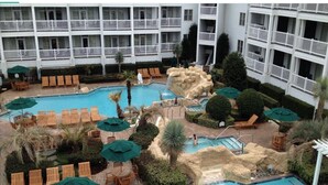 A hotel pool area with multiple lounge chairs and umbrellas, surrounded by a building with balconies. - Turtle Cay - Key-West Style 2Bedrm 2.5bath Full Week Oceanview July 10-17 (Virginia Beach)