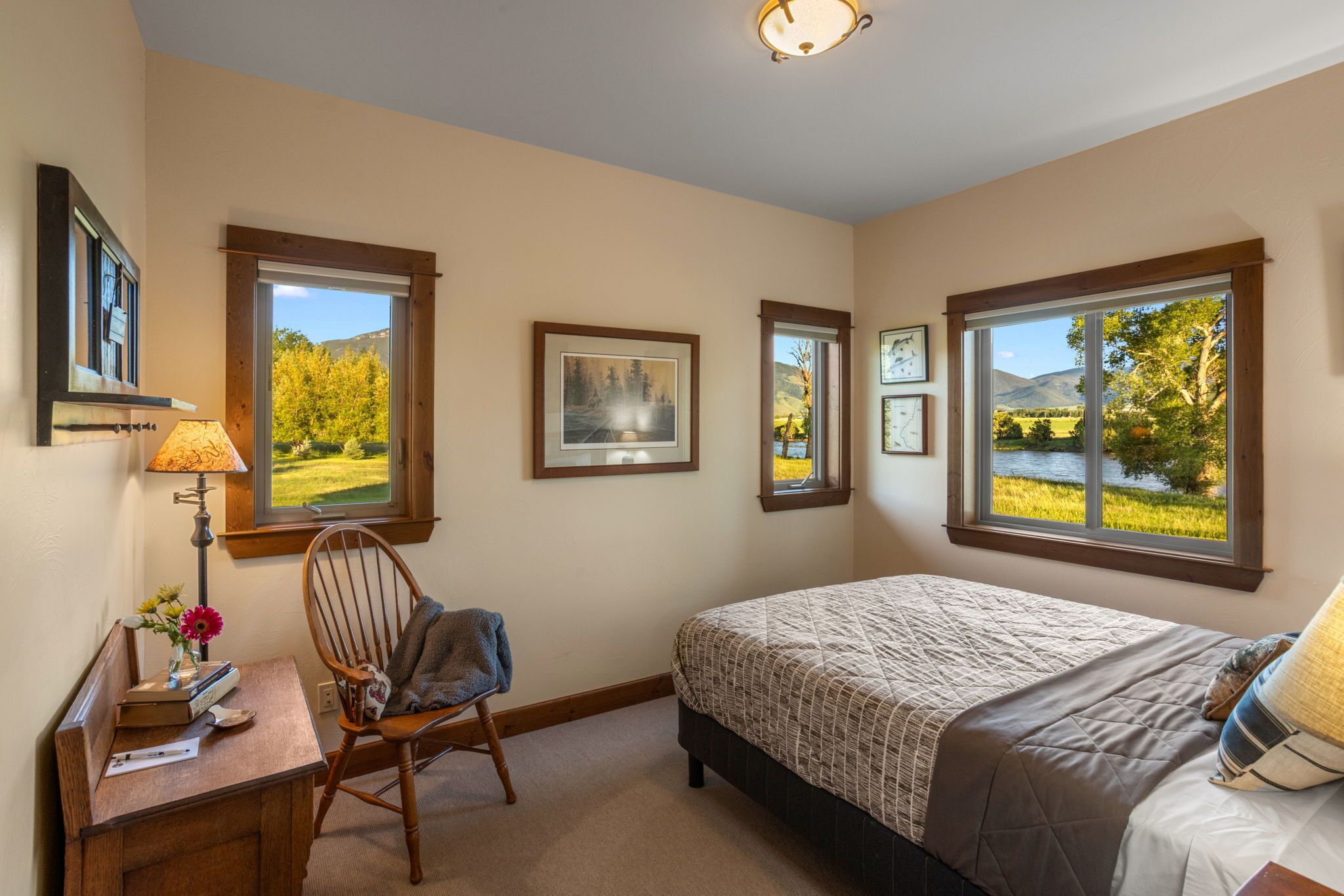 A bedroom with a large bed, a desk with a lamp, a chair, and multiple windows offering views of a lake and trees.