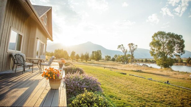 A wooden deck with outdoor furniture, a flower pot, and a view of a lake and mountains. - Tranquil Home Along the Yellowstone River (Emigrant)
