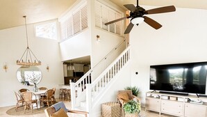 A spacious living room with a high ceiling, a ceiling fan, a large flat-screen TV, a dining area with wicker furniture, and a staircase leading to an upper level. - 3BR/3BA Villa: Central A/C! Short Walk To Beach! (Koloa)