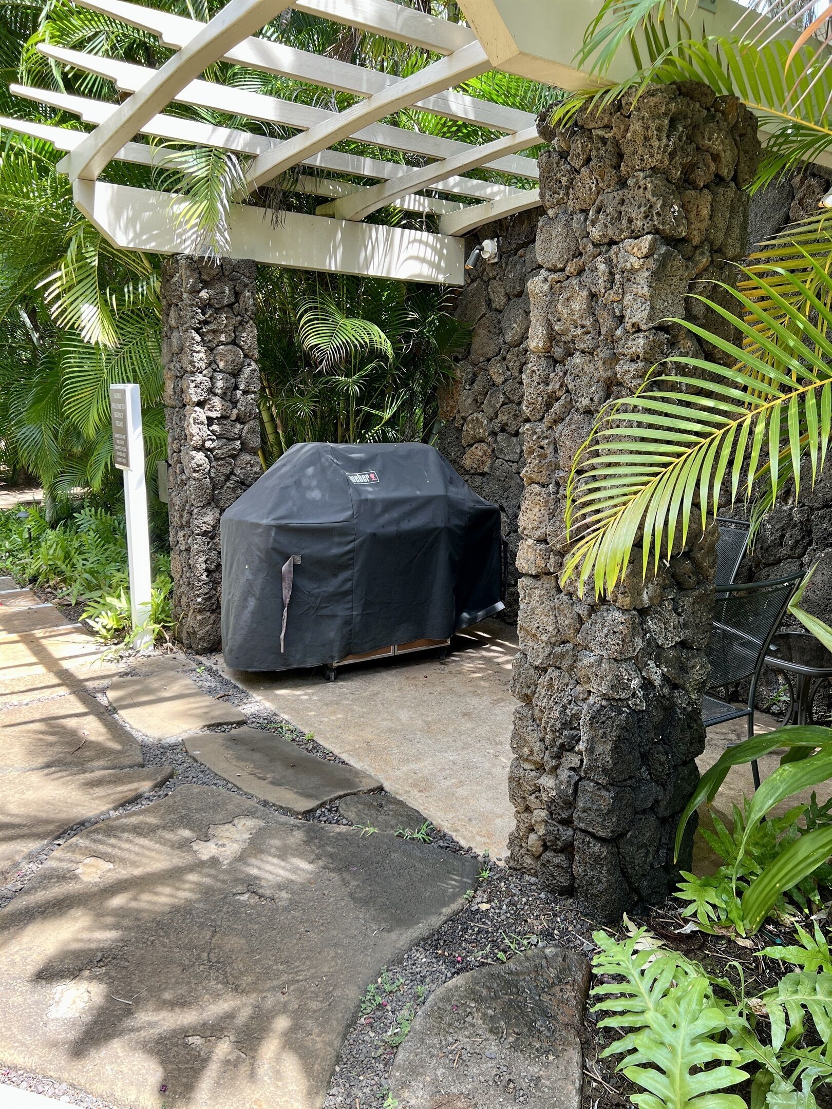 Ein überdachter Grillbereich mit Steinwand, weißer Pergola und üppiger tropischer Bepflanzung.