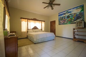 A bedroom with a bed, a ceiling fan, a painting, a television, and a wooden cabinet. - Rainbow Hill (Negril)