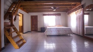 A bedroom with a wooden staircase, a bed with floral bedding, a ceiling fan, and a window with curtains. - Rainbow Hill (Negril)