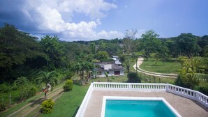 A pool with a white fence, surrounded by lush greenery and a house in the background. - Rainbow Hill (Negril)