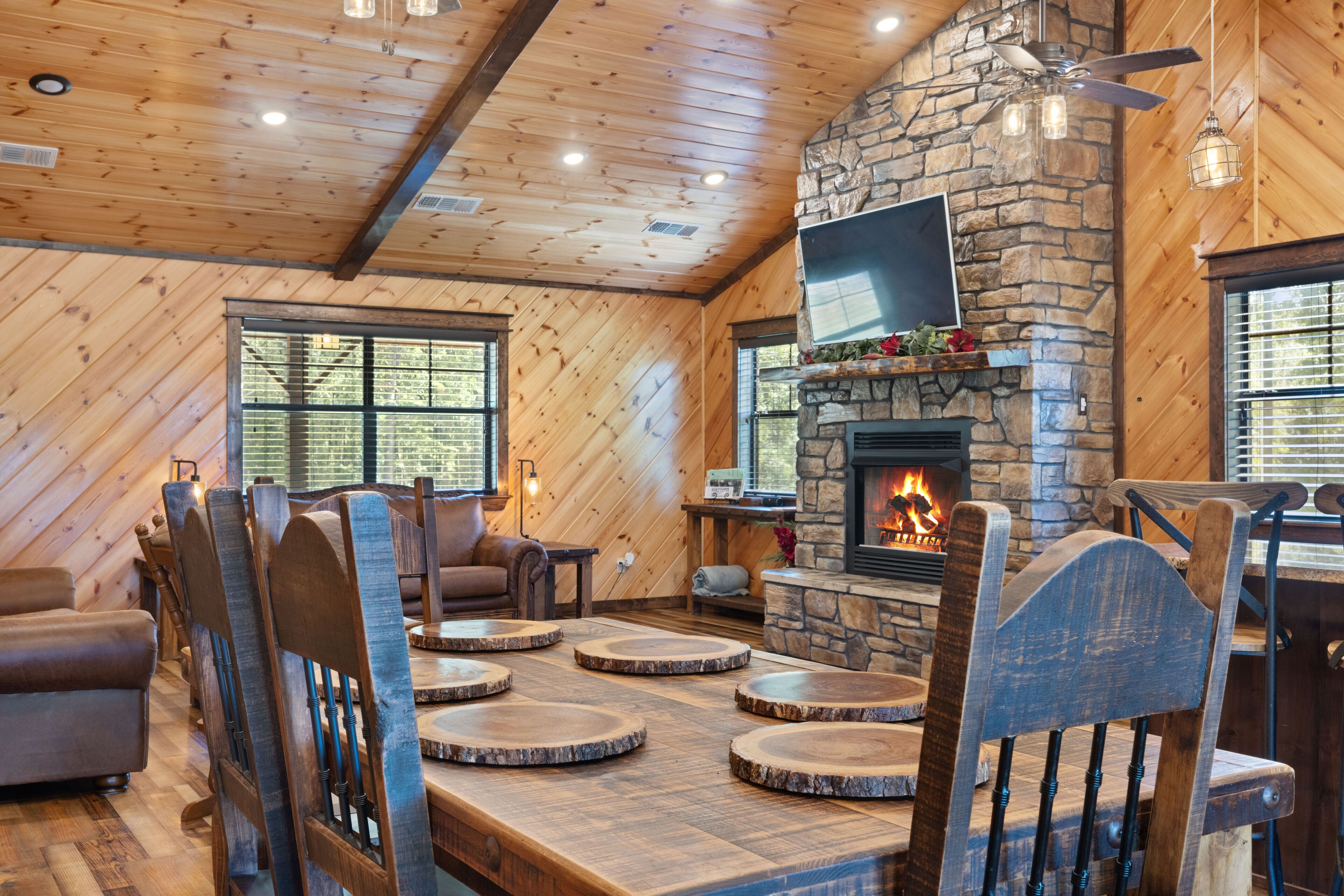 A cozy cabin interior with a stone fireplace, wooden walls, and a dining area with round wooden tables and chairs.