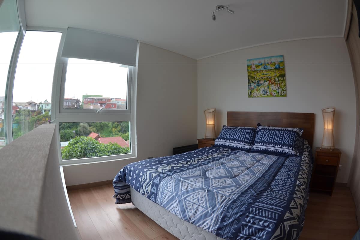 Um quarto com uma janela grande, uma cama com roupa de cama estampada, uma mesinha de cabeceira com abajur e um quadro colorido na parede.