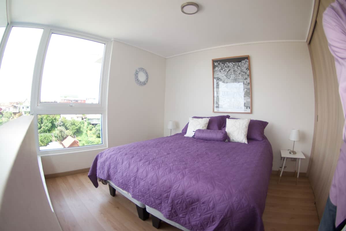 Quarto com cama grande, janela, mesa de cabeceira e quadro na parede.
